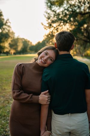 A couple embraces in a warm sunset glow on a grassy field with trees in the background.