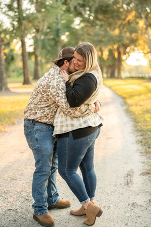 Couple sharing an intimate moment on a sunlit gravel path surrounded by trees.