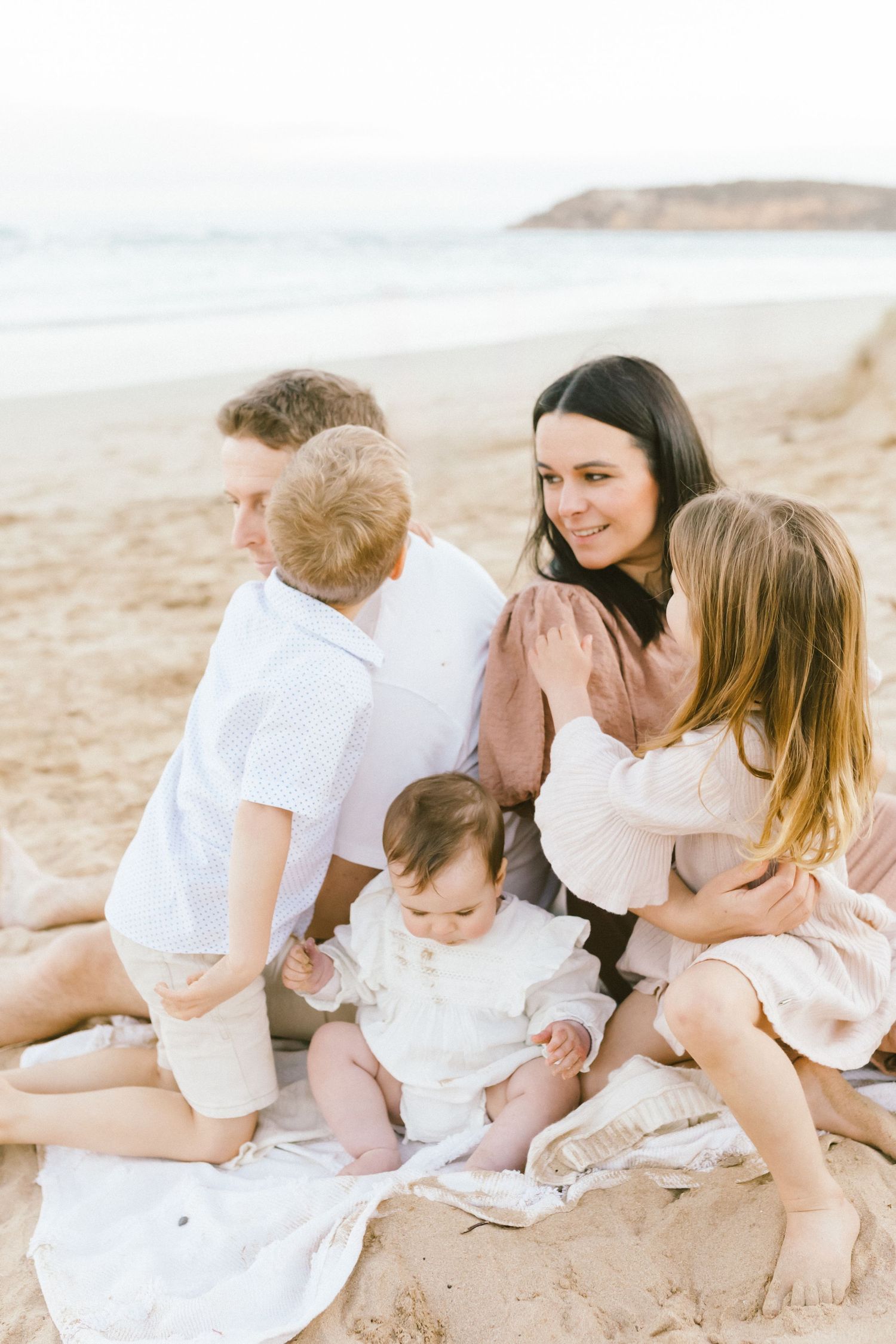 Family enjoying tender moments together on a sandy beach in matching white and pink outfits.