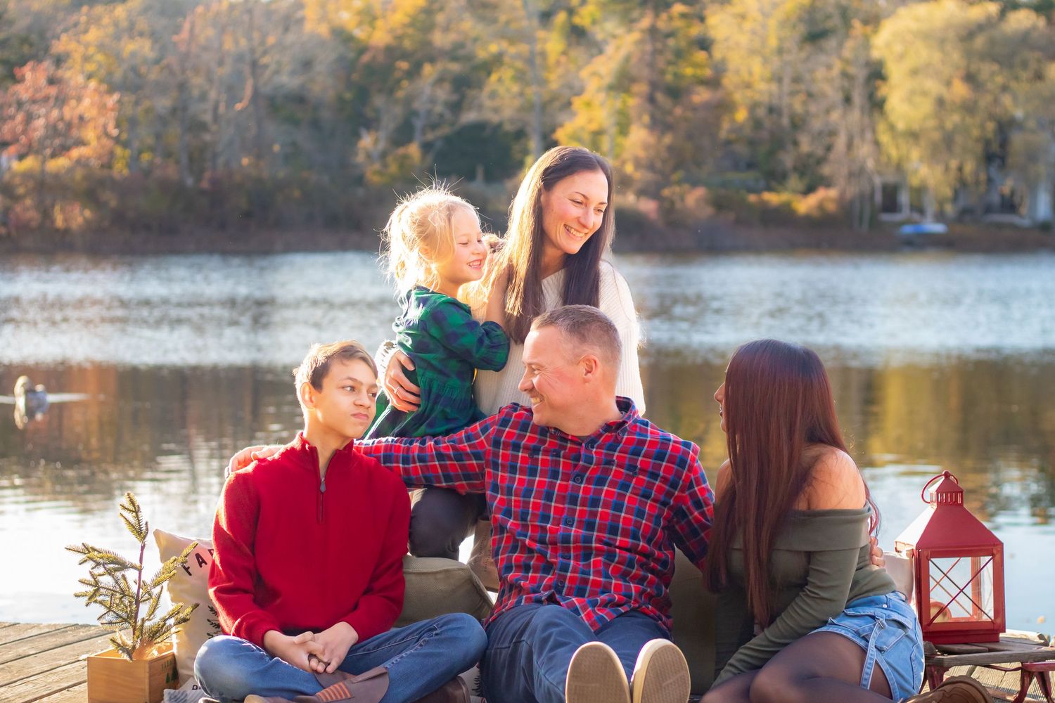 People holding Christmas-themed wooden signs while sitting outdoors near a lake at sunset.