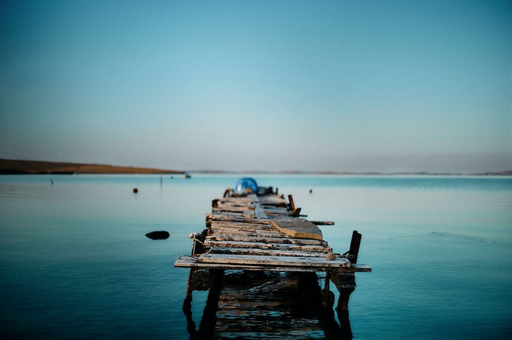 Weathered wooden pier stretches into calm blue waters with distant mountains creating a serene seascape.