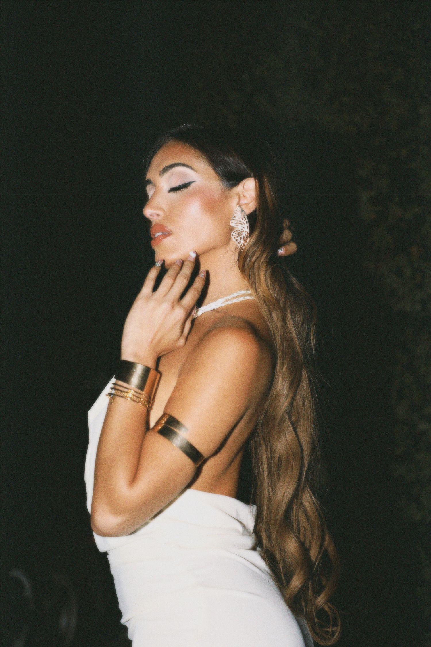 Woman in white dress poses dramatically against dark background with sparkling jewelry and flowing hair.