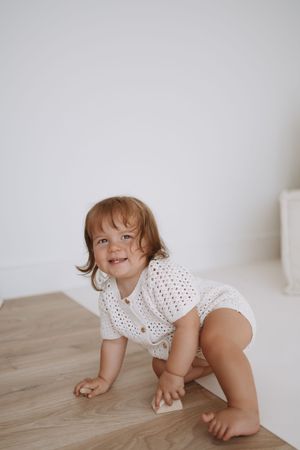 A small child in a polka dot outfit crawls and plays on a light wooden floor against a white wall.
