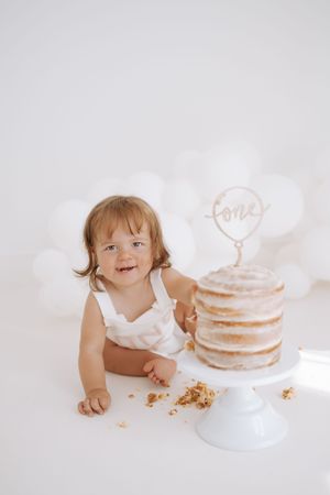 A small child in white sits beside a minimalist naked cake with crumbs on a white backdrop during a cake smash photo session.