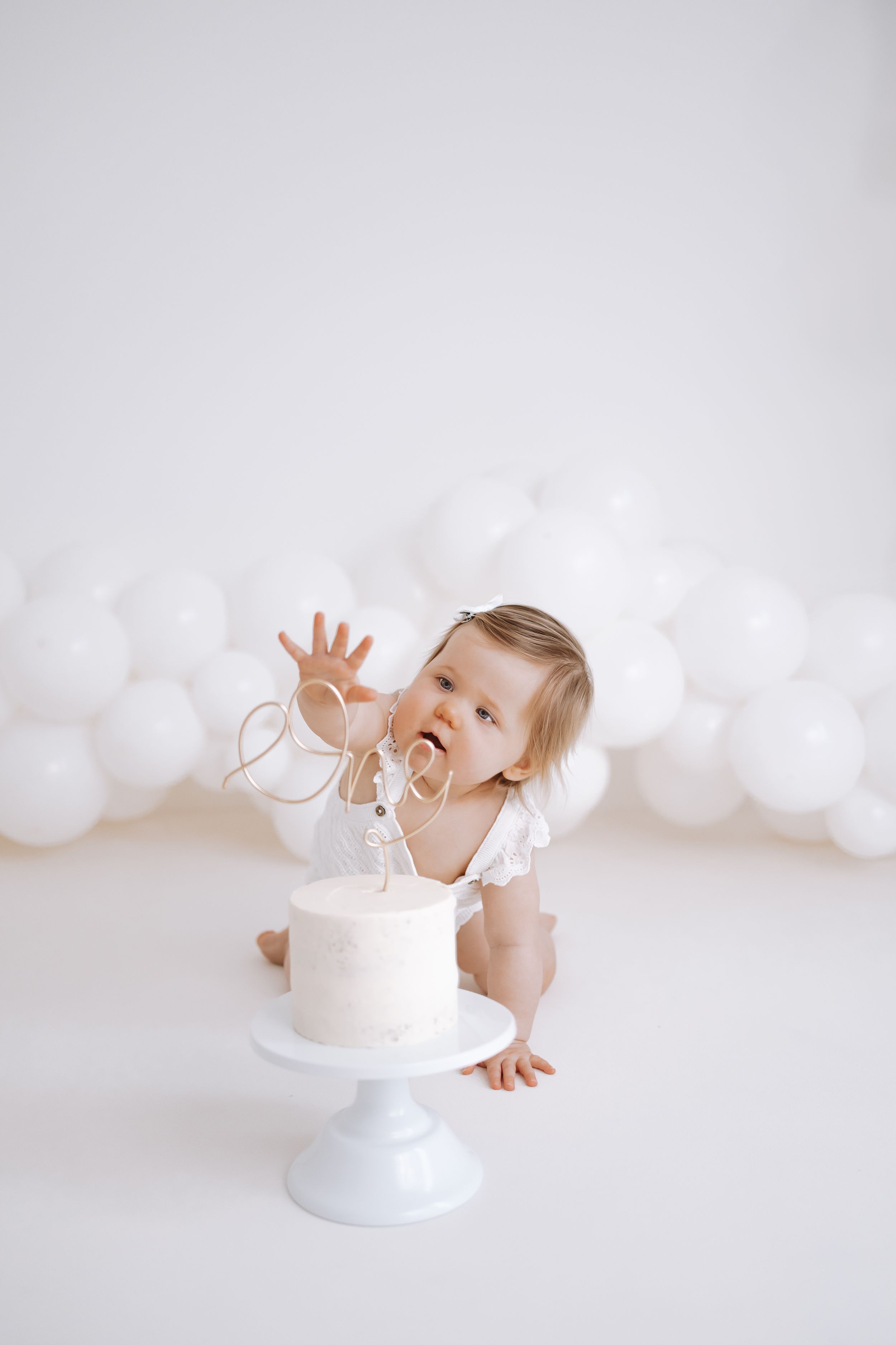 A baby in white dress reaches for a simple white birthday cake against a soft white background in a minimalist style.