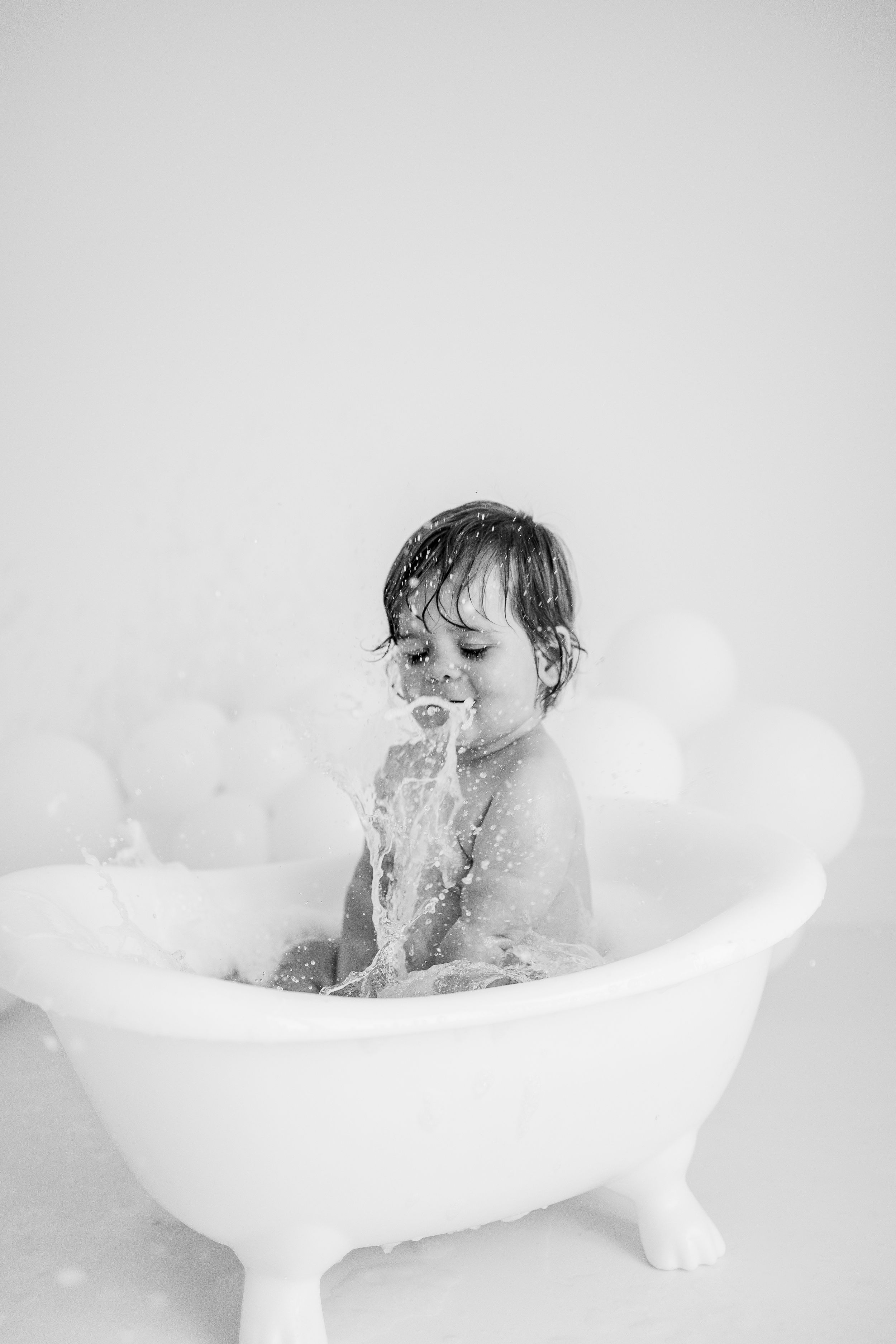 A child splashes and plays in a white bathtub filled with soapy bubbles in this dreamy black and white photograph.