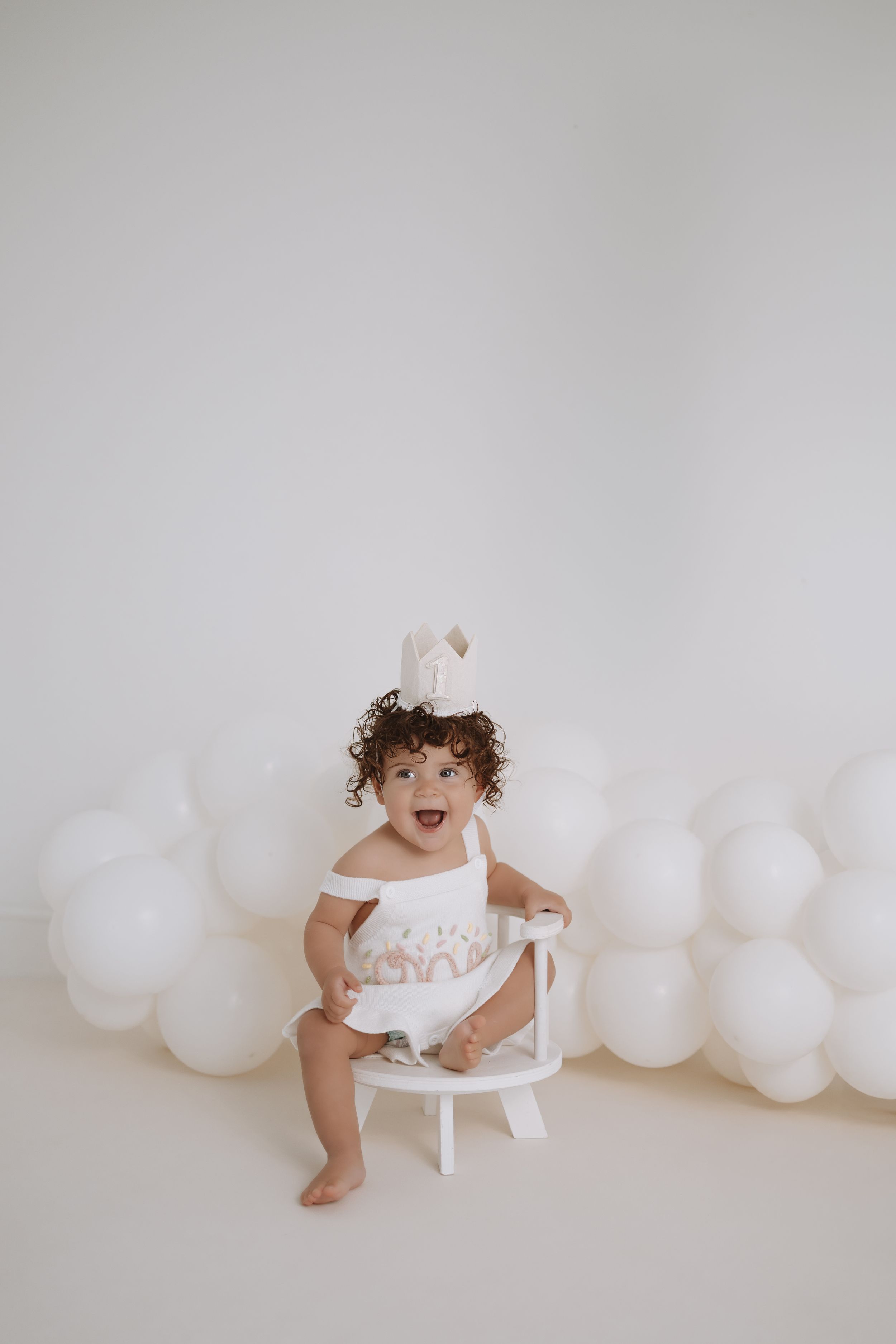 A toddler in a white dress sits and plays on a white chair against a backdrop decorated with white balloons.