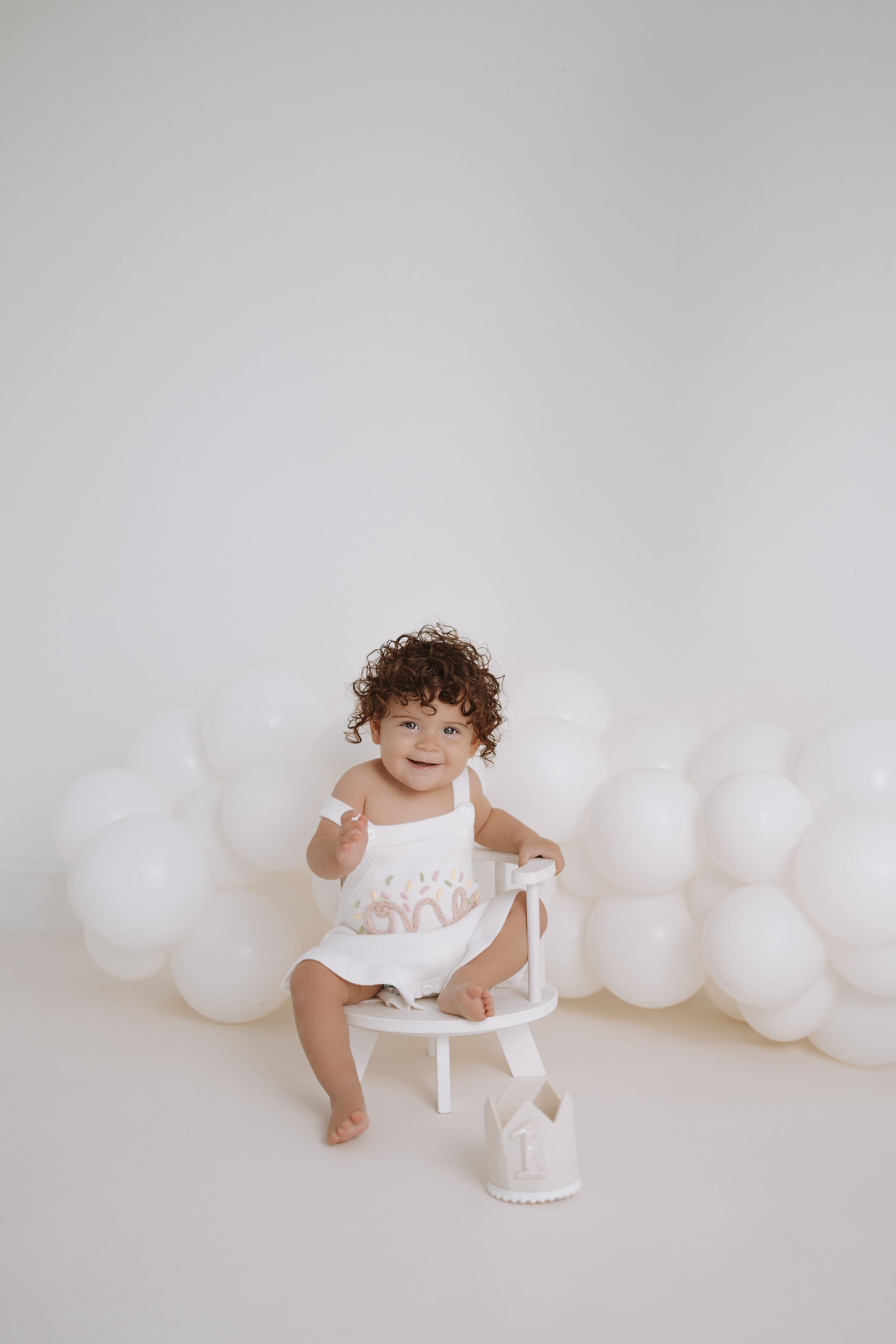 A baby in a white dress sits on a small chair against a white background with white balloons.
