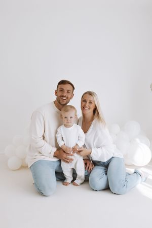 Family in matching white shirts and light blue jeans sitting together on white background with stuffed animals.