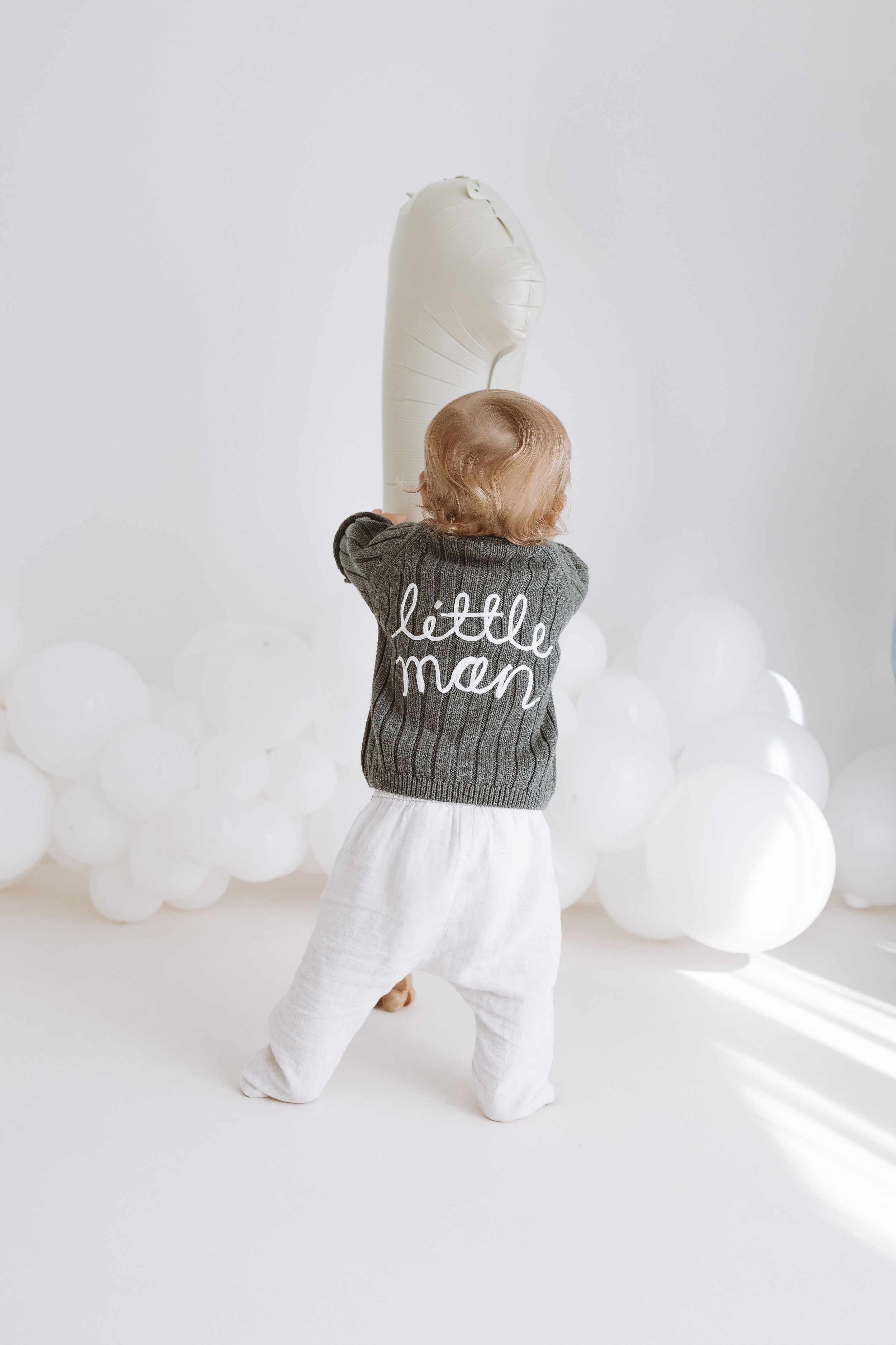 Baby in white onesie and gray knit beanie crawls on white floor in minimalist setting.