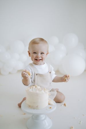 A white cake smash photo session showing a baby in white outfit playing with a small cake on a pedestal.