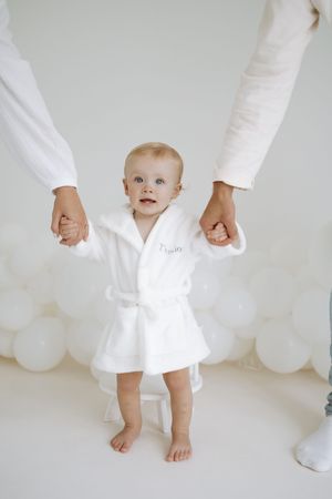 Baby wearing white robe standing with adult hands visible against white backdrop with balloons.
