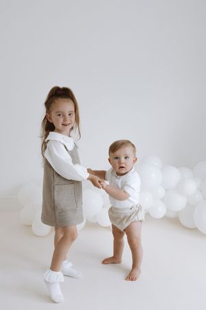 Two children in white and gray outfits play together against a white background with balloon decorations.