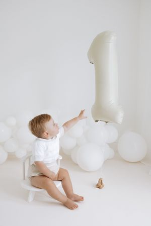 Baby in white outfit crawls and plays with white balloons in minimalist white studio setting.