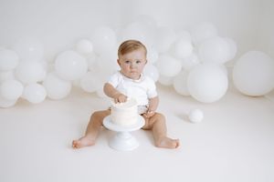Baby wearing white clothing sits on floor surrounded by white balloon decorations in minimalist photo setting.