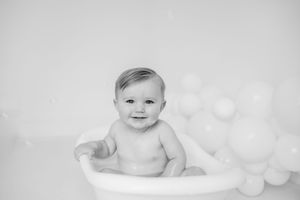 Black and white portrait of a baby sitting in a bathtub with soft bokeh lights in the background.