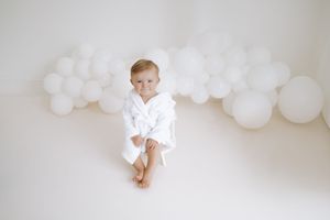 Small child wearing white robe sitting against white wall decorated with balloon garland.