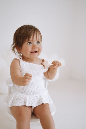 A happy baby wearing a white smocked dress sits on a white chair against a light gray background.