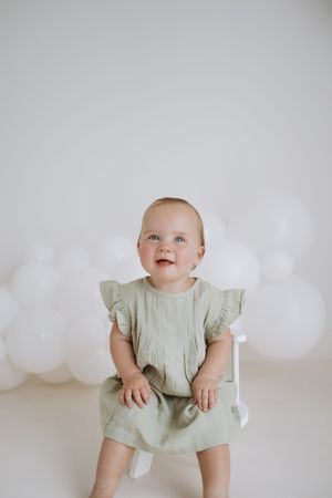 Baby in sage green flutter sleeve dress sits against white background with balloon decorations.