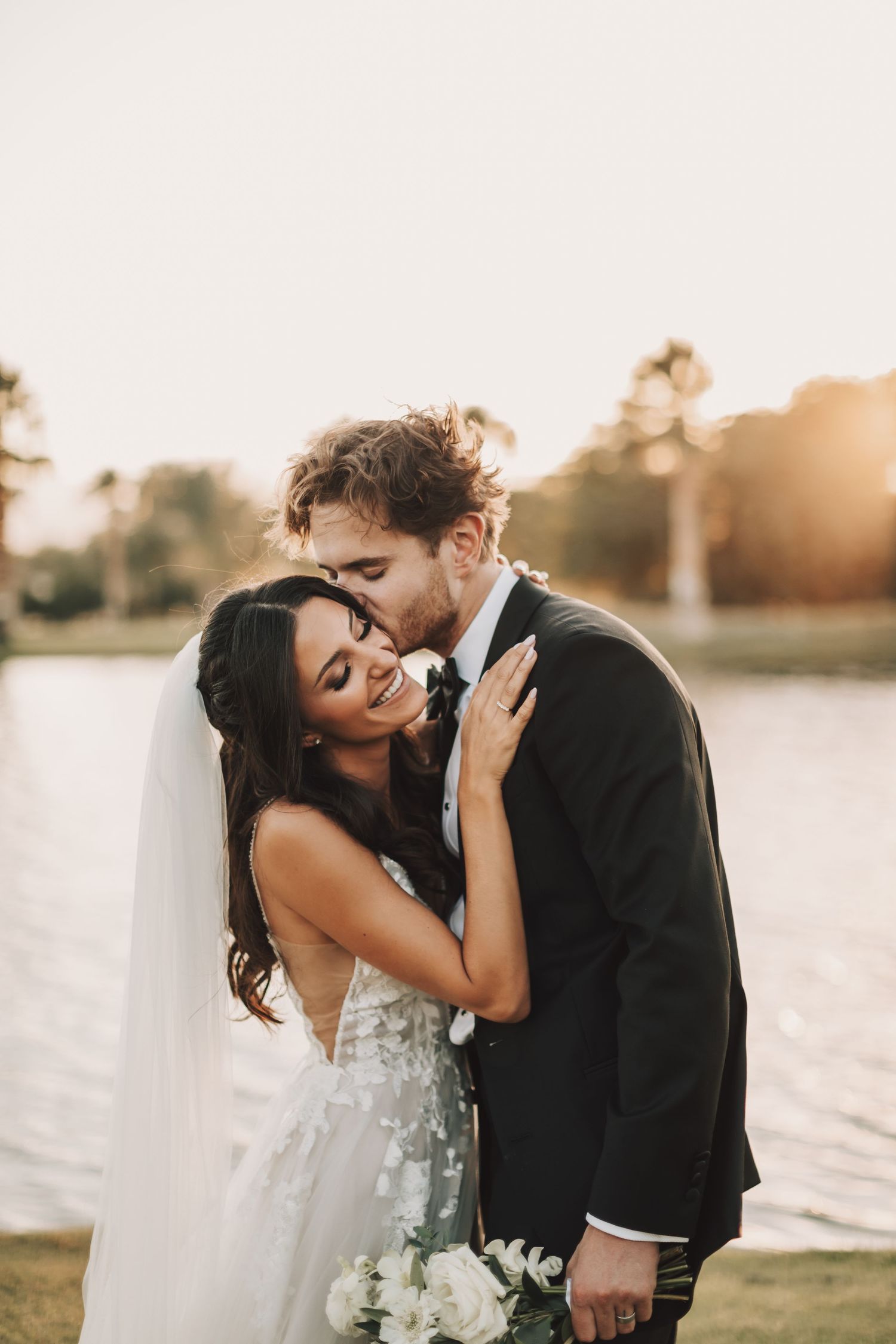 A romantic wedding couple shares an intimate moment at sunset by a lake, bride wearing a flowing embellished gown.
