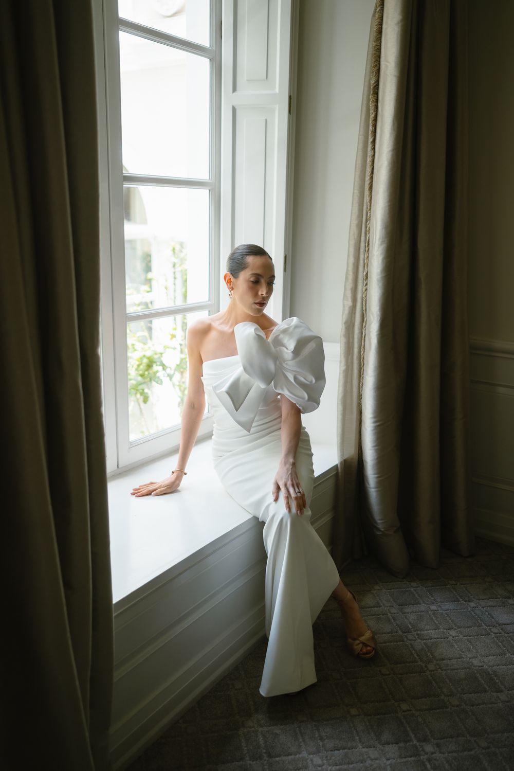 An elegant bridal portrait by a window featuring a modern white structured wedding dress.