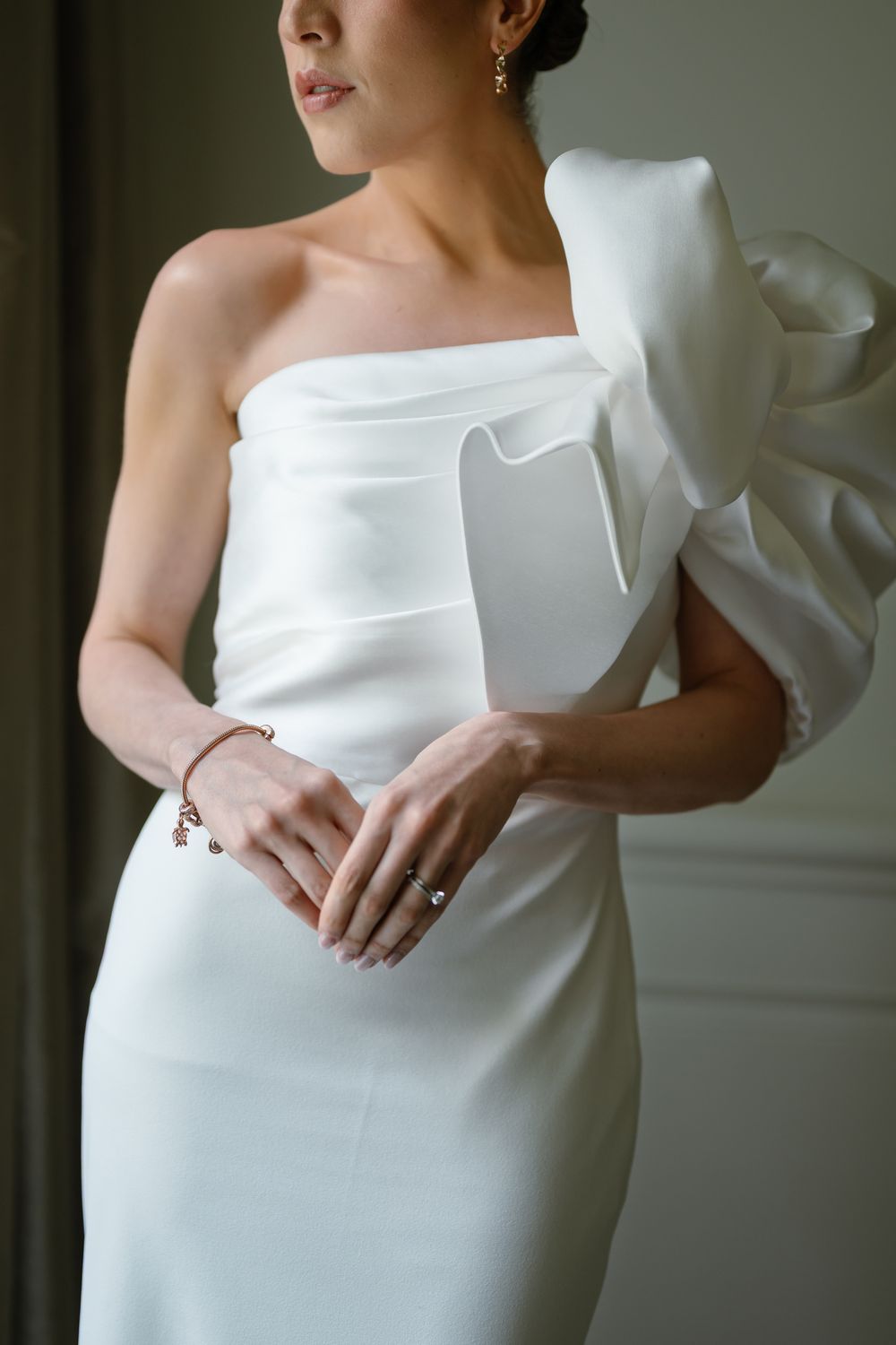 Bride in elegant white one-shoulder gown with dramatic bow detail, wearing delicate jewelry, hands clasped gracefully.