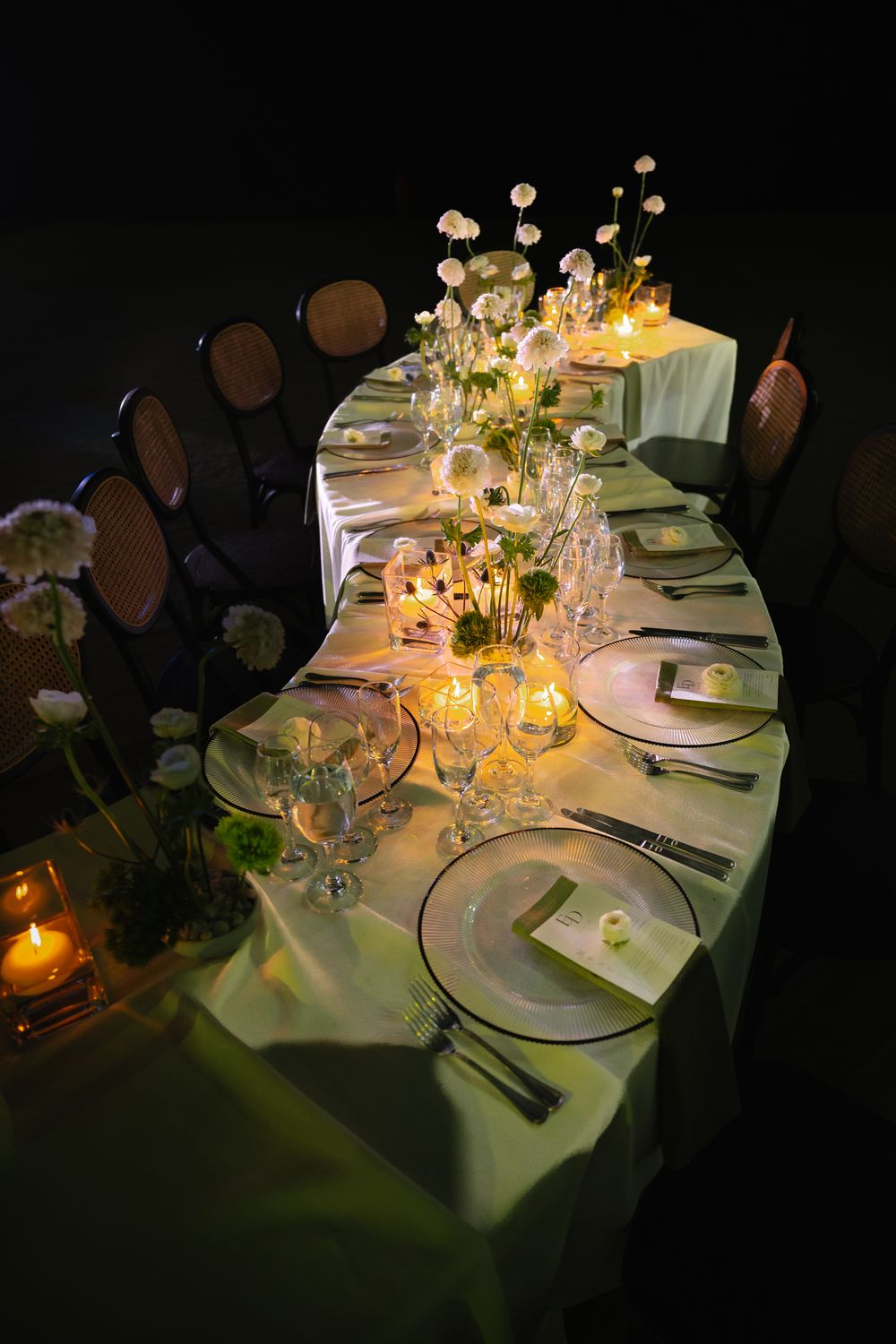Elegant candlelit dinner table with white floral centerpieces, green linens, and formal place settings in a dark setting.