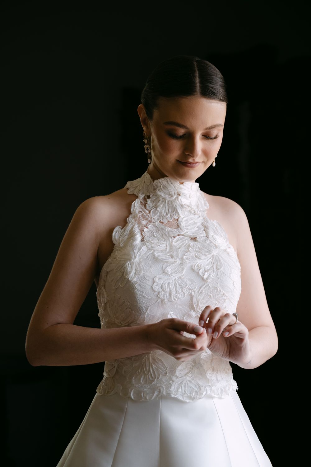 Bride in white lace halter wedding gown adjusting her ring, elegant dark background portrait.