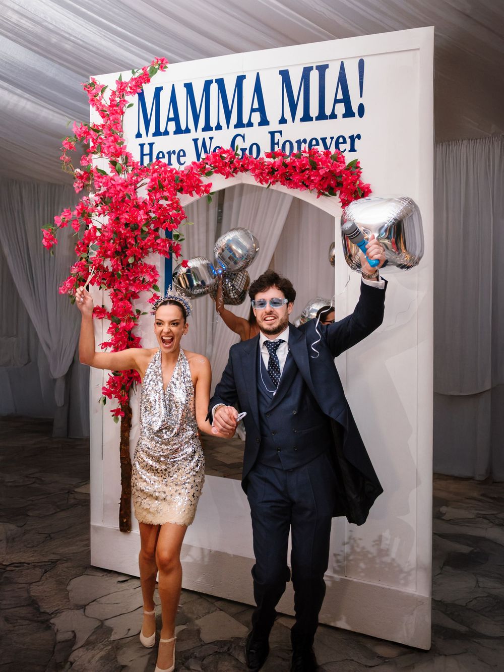 A couple in formal attire dancing and posing in a white room with pink floral decorations and Mamma Mia themed signs.