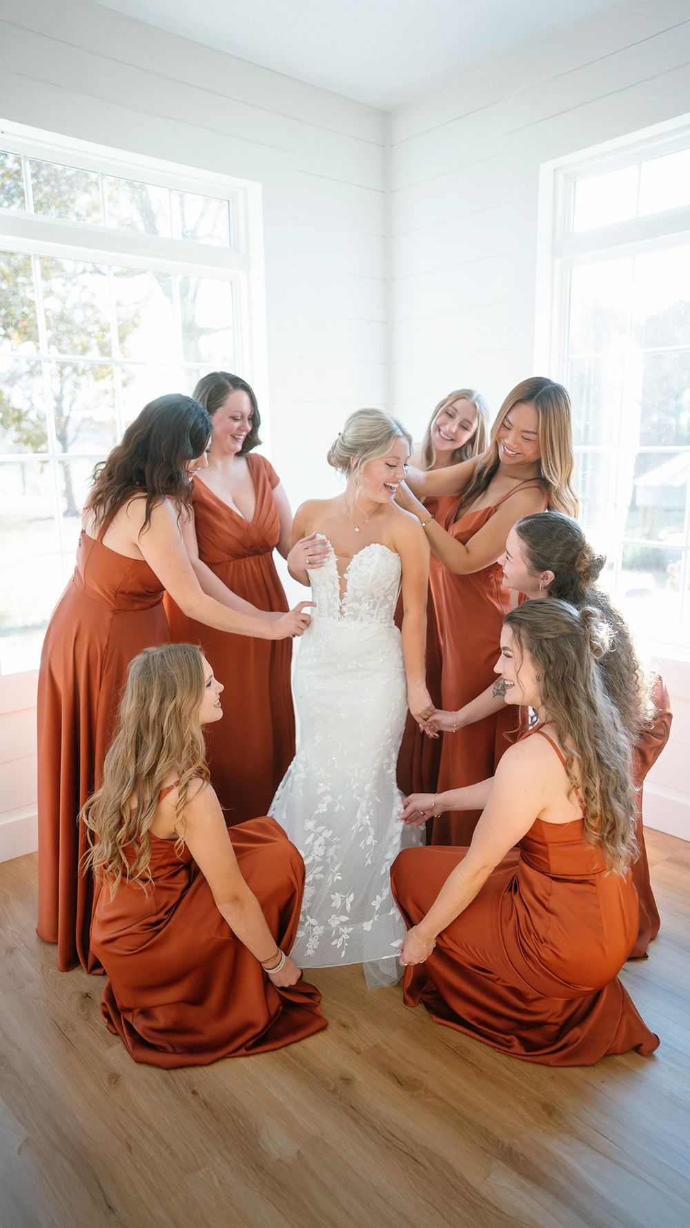 Bride in white lace gown surrounded by bridesmaids in rust orange dresses in bright white room.