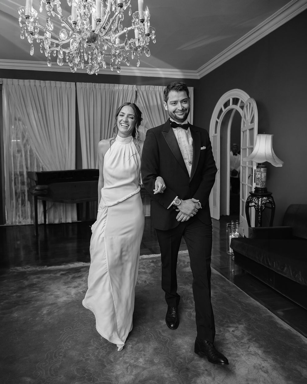 Elegant couple in formal attire, woman in white gown and man in tuxedo, posing in a grand room with crystal chandelier.