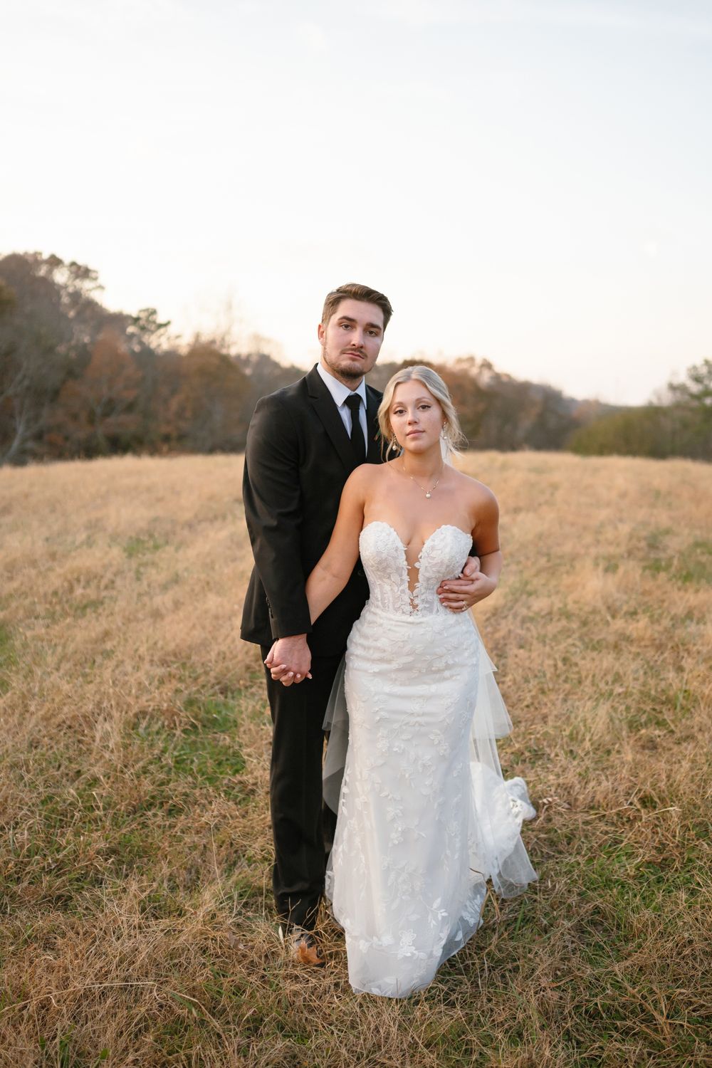 A bride in a lace gown and groom in a black suit pose in a golden field at sunset.