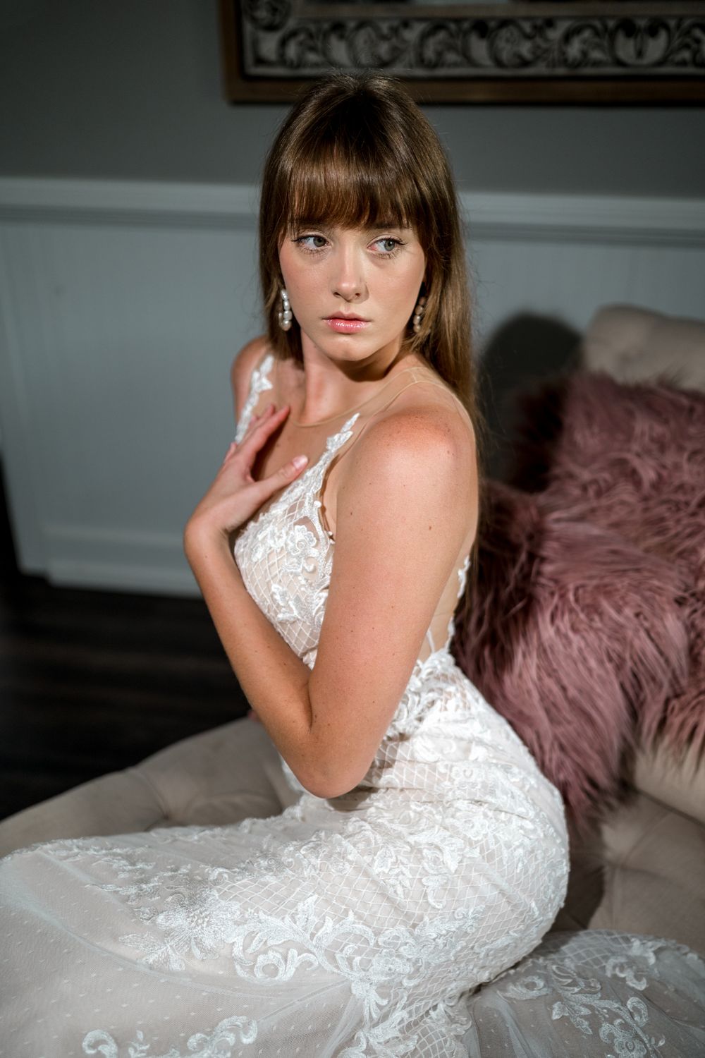 Elegant brunette bride in fitted lace wedding gown sitting gracefully on a vintage sofa indoors.