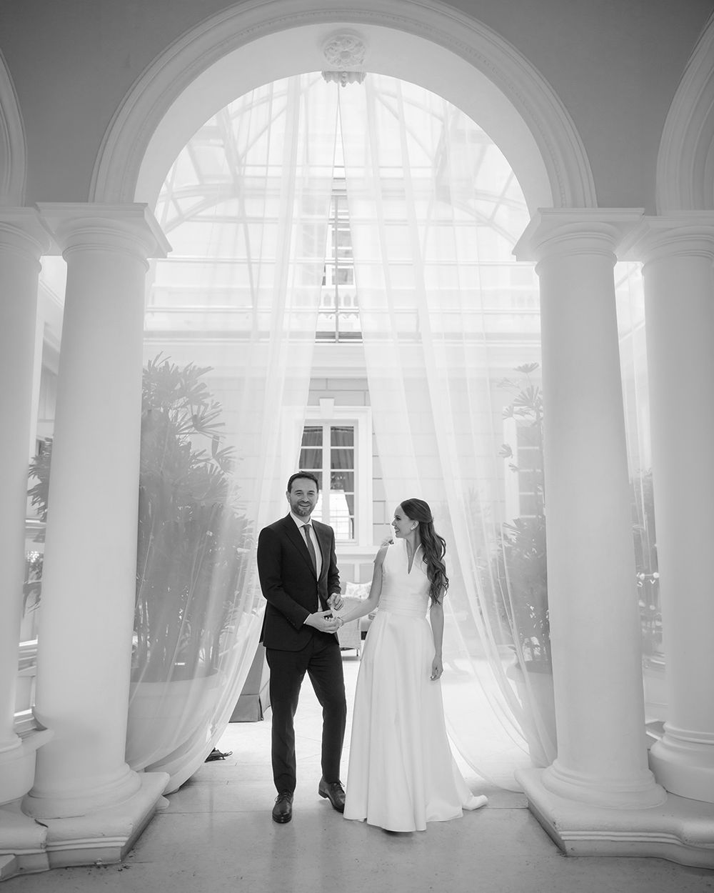 Black and white wedding photo of bride and groom holding hands under elegant arched colonnade with flowing white drapes.