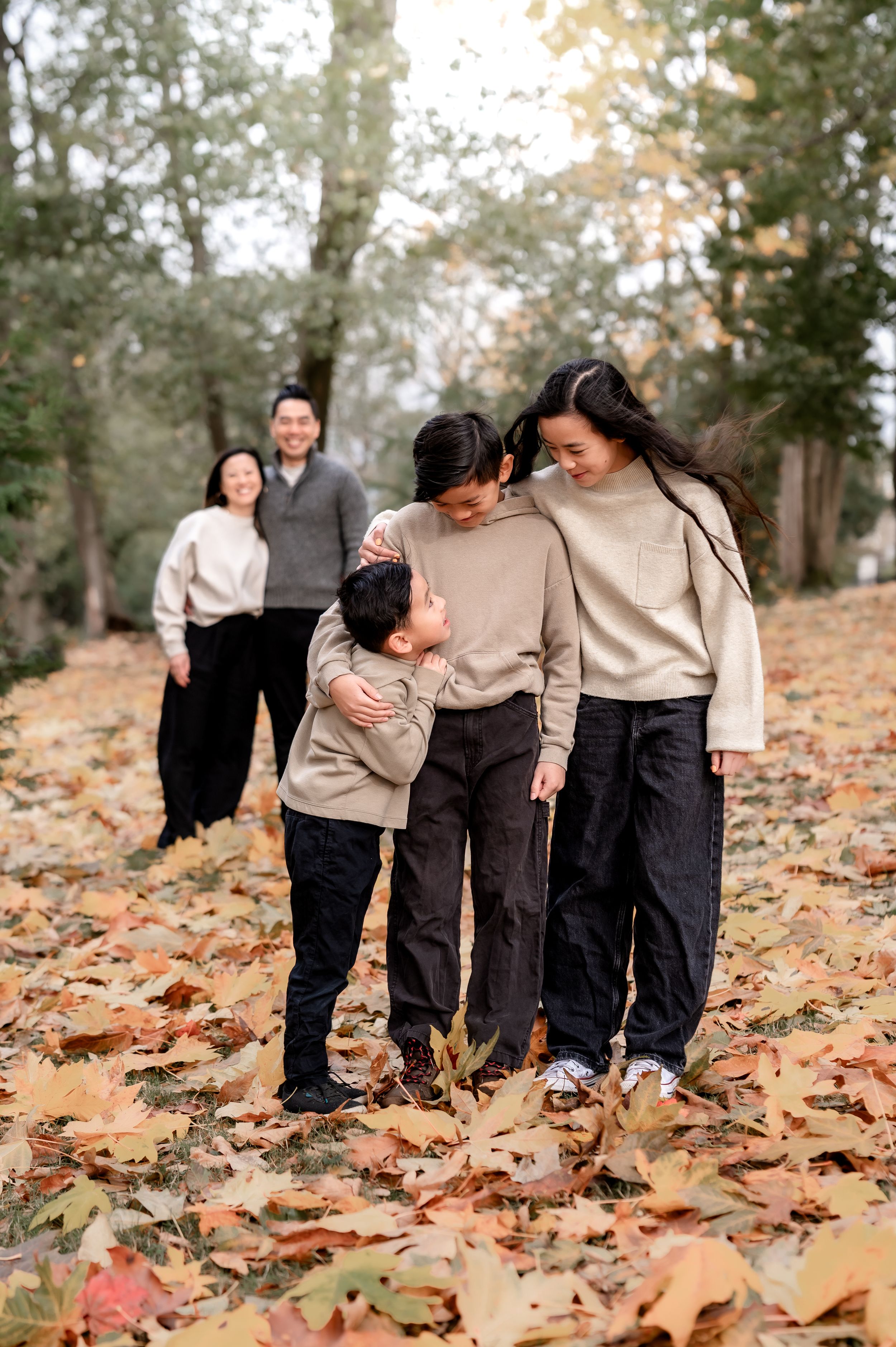 A family wearing coordinated beige sweaters and dark pants walks together through autumn leaves in a wooded setting.