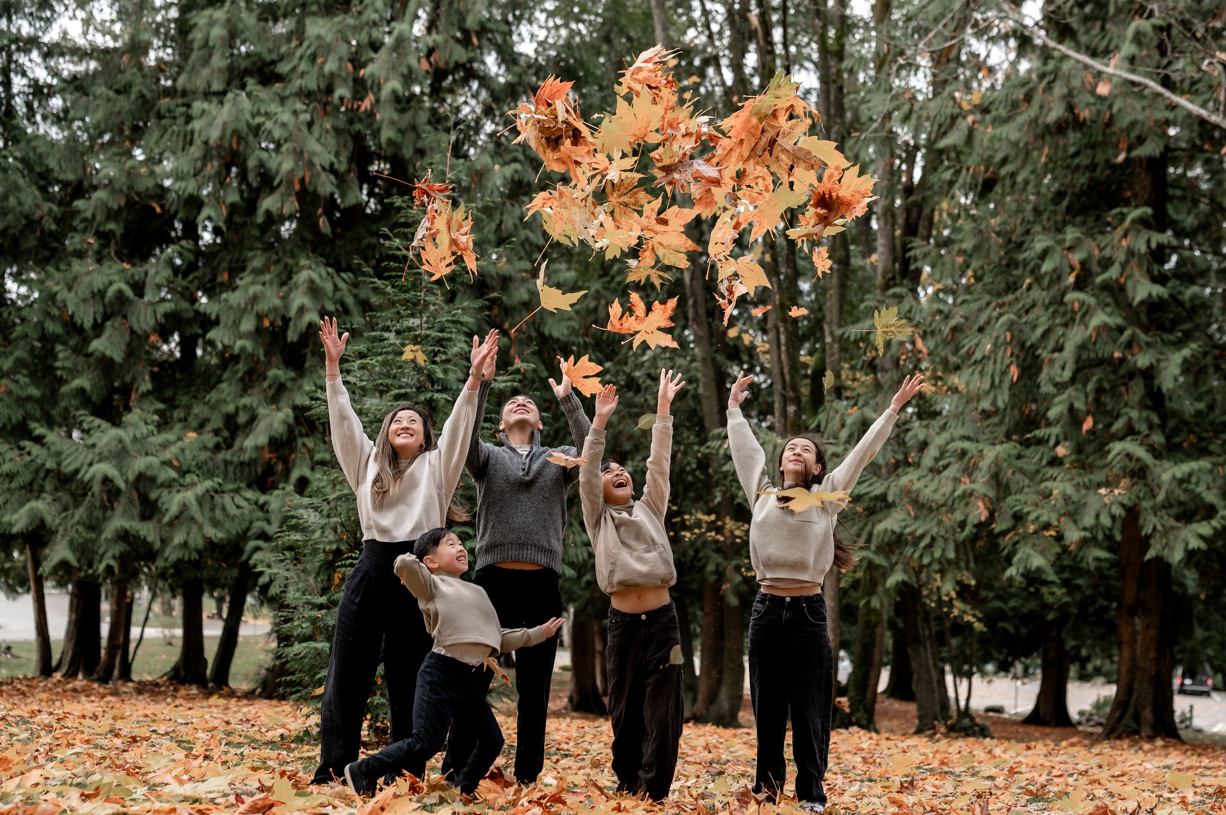 A group celebrates joyfully in an outdoor autumn setting with falling orange leaves and tall trees in the background.