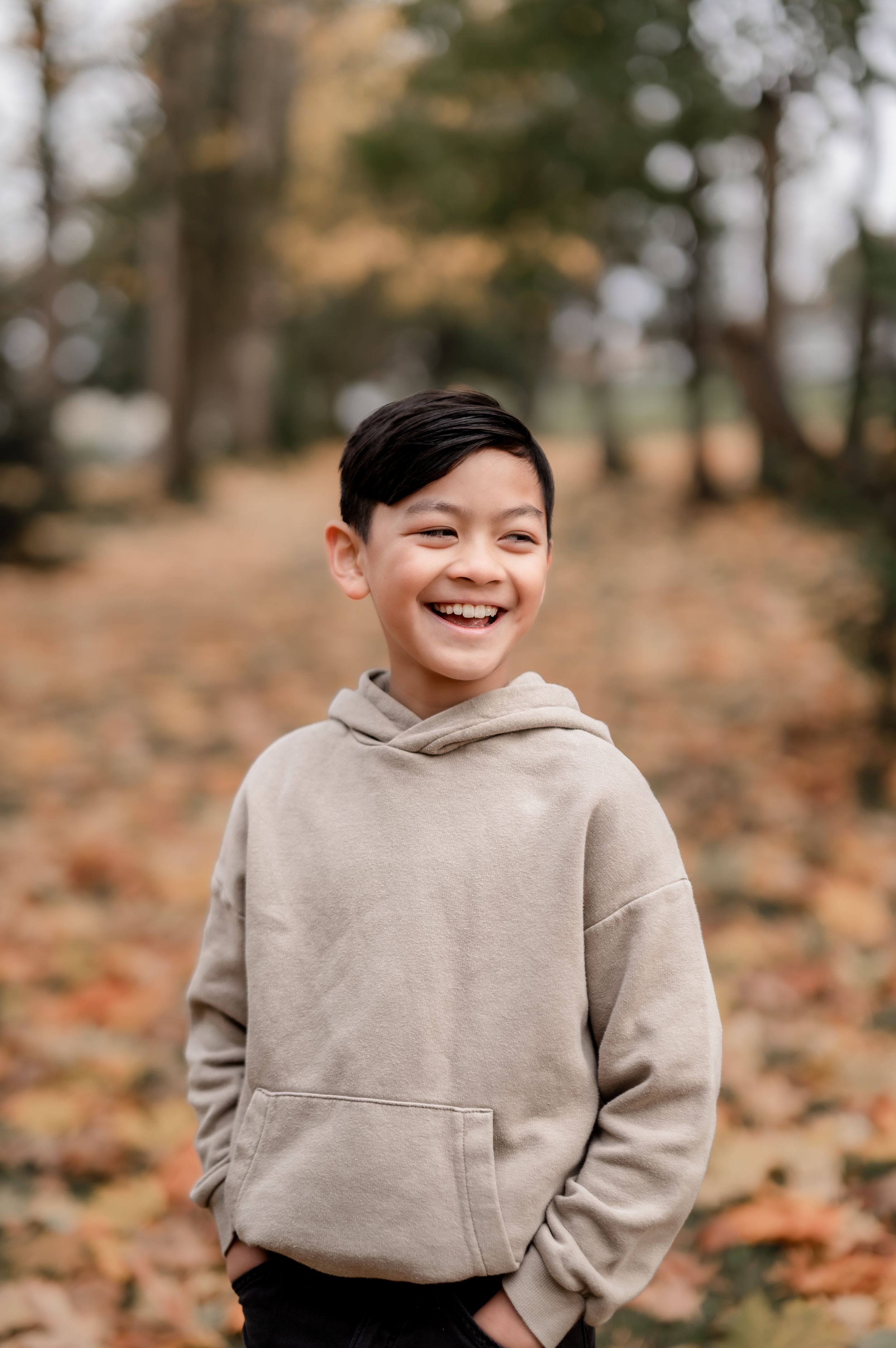 Young child wearing beige hoodie smiling and laughing while sitting among autumn leaves outdoors.