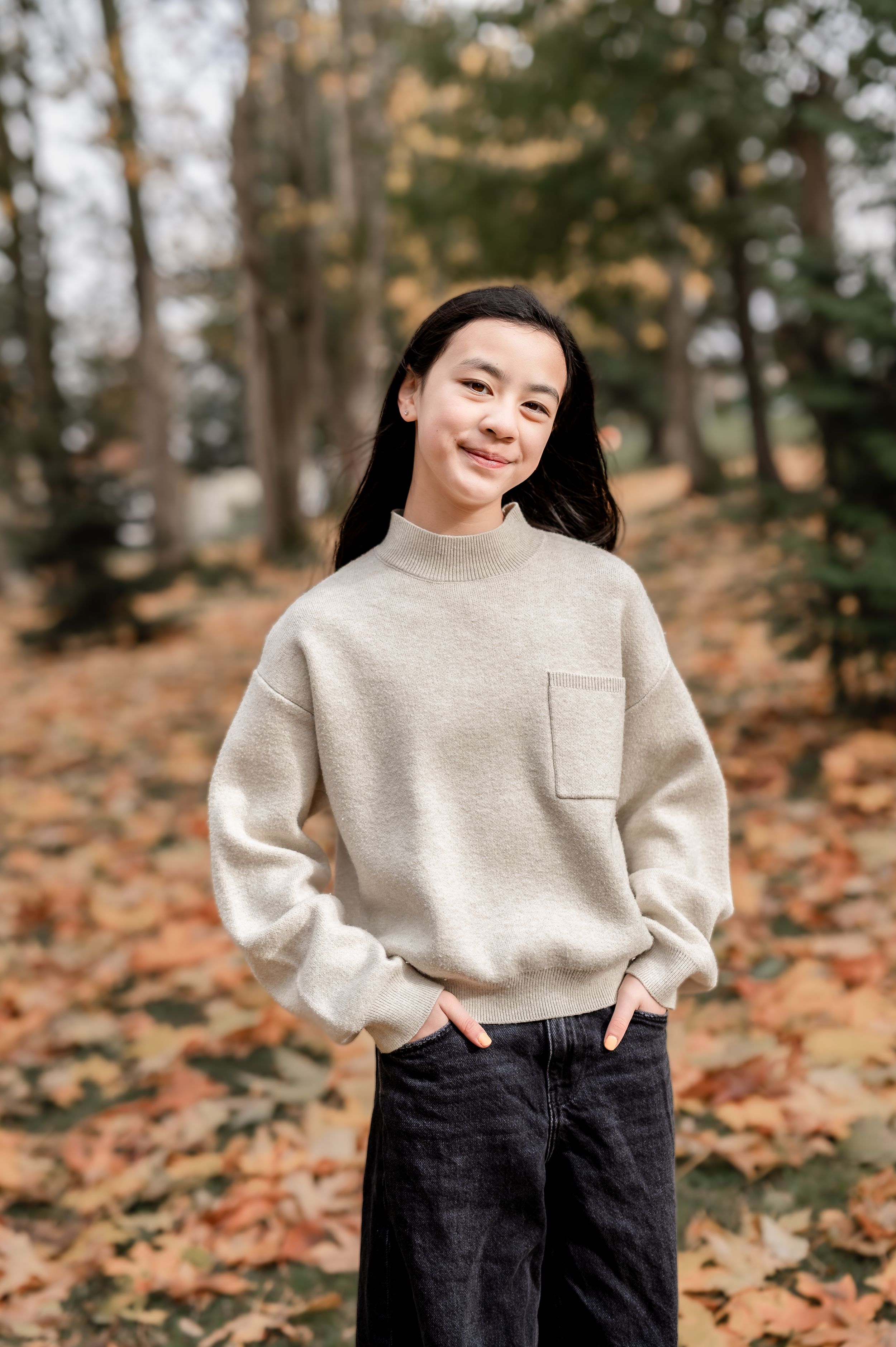 Person in cream sweater and black pants poses outdoors among fallen autumn leaves in wooded setting.