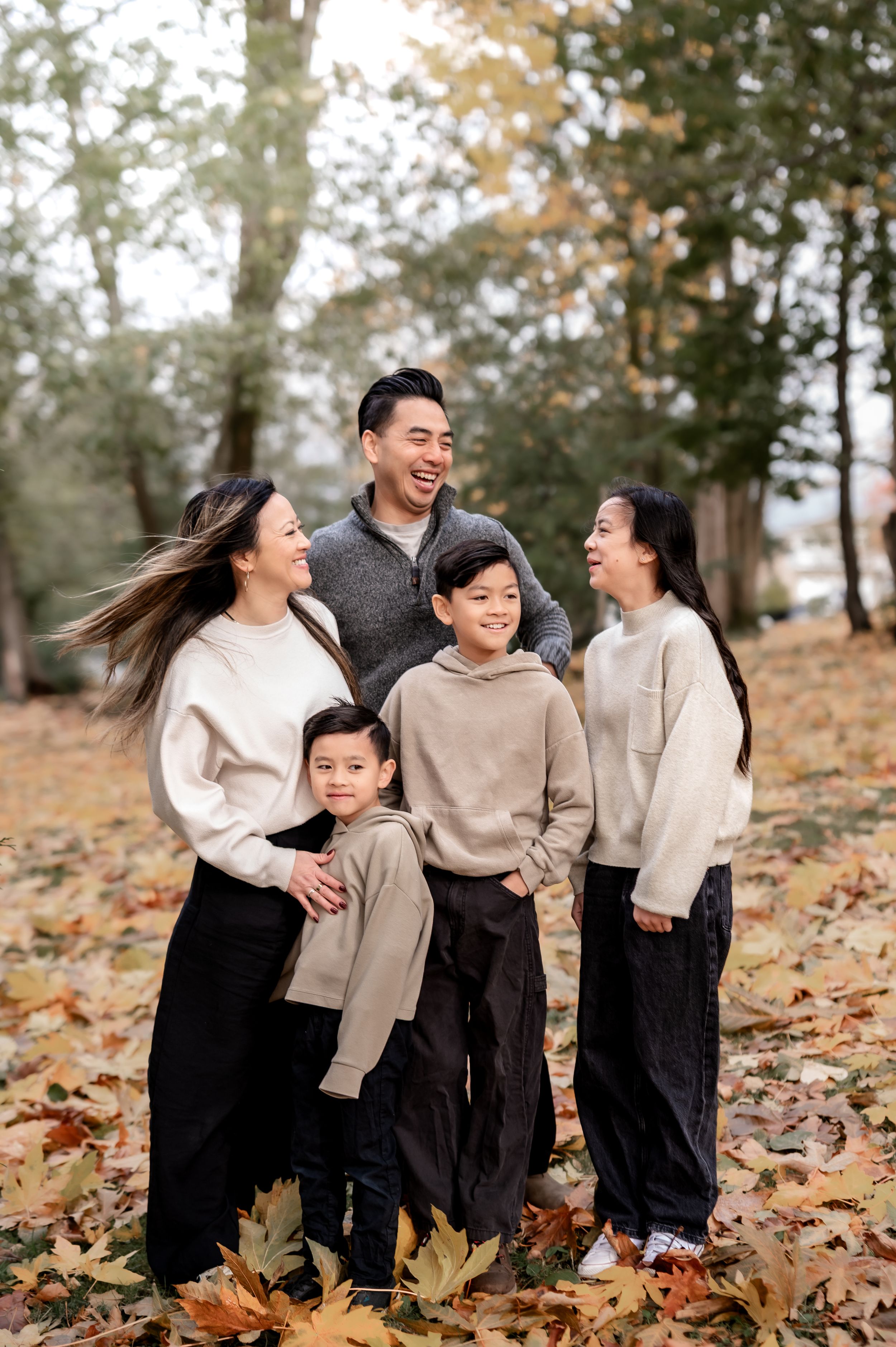 Family wearing coordinated beige and black outfits laughs together while posing among fallen autumn leaves in a park.