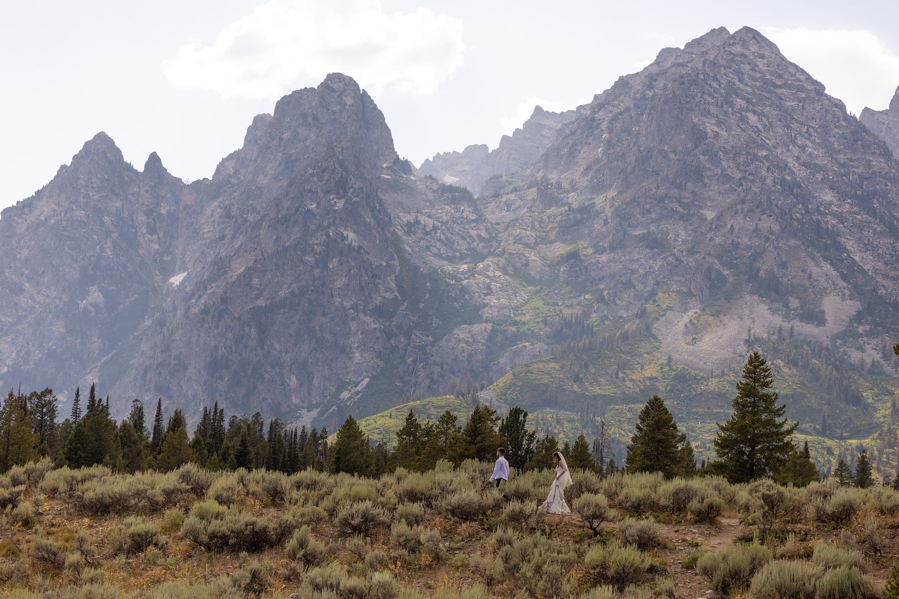 Rugged mountain peaks rise above evergreen trees in Grand Teton National Park on a cloudy day.