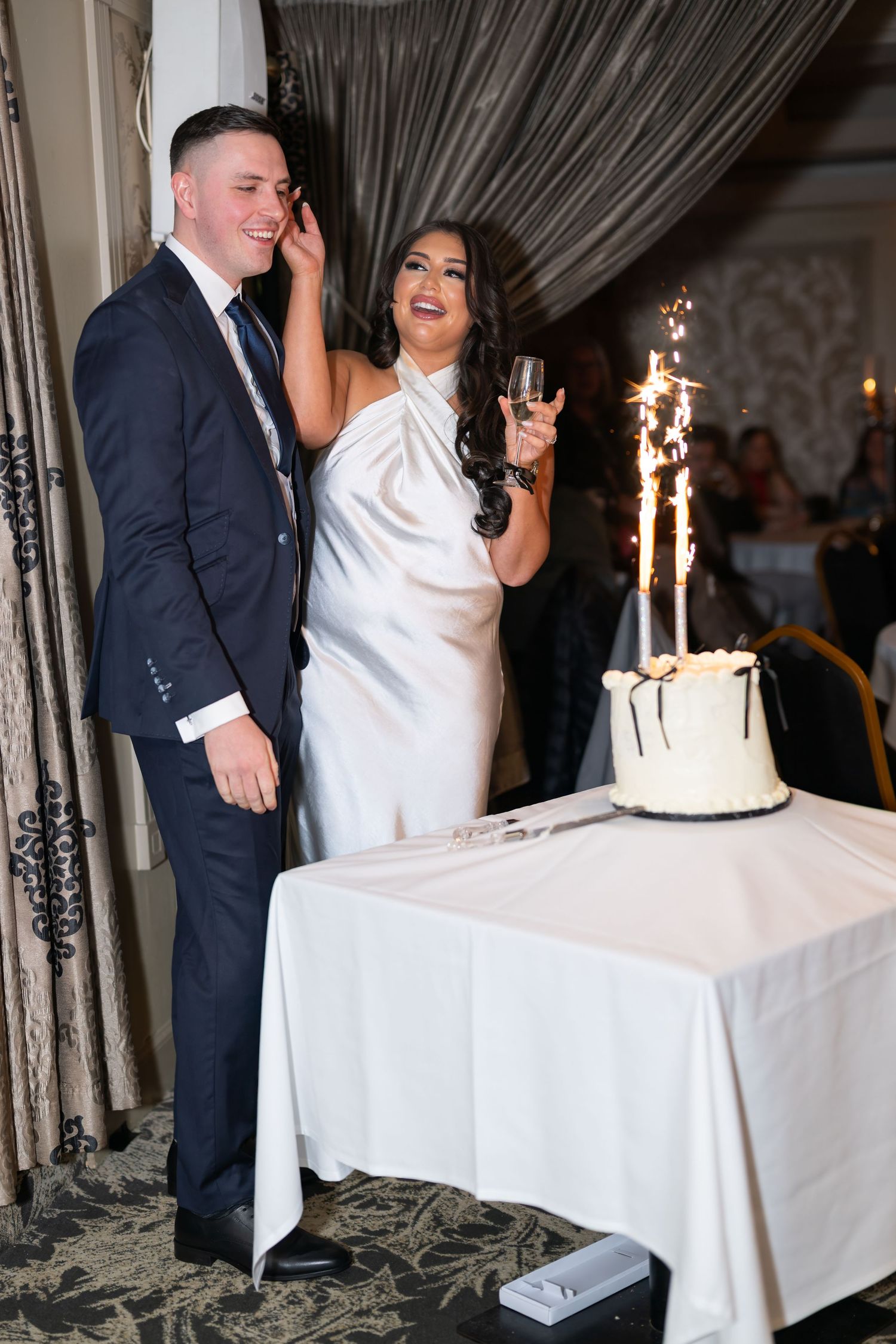 A wedding reception scene with a tiered white cake with sparkler candles on a table draped in white linens.