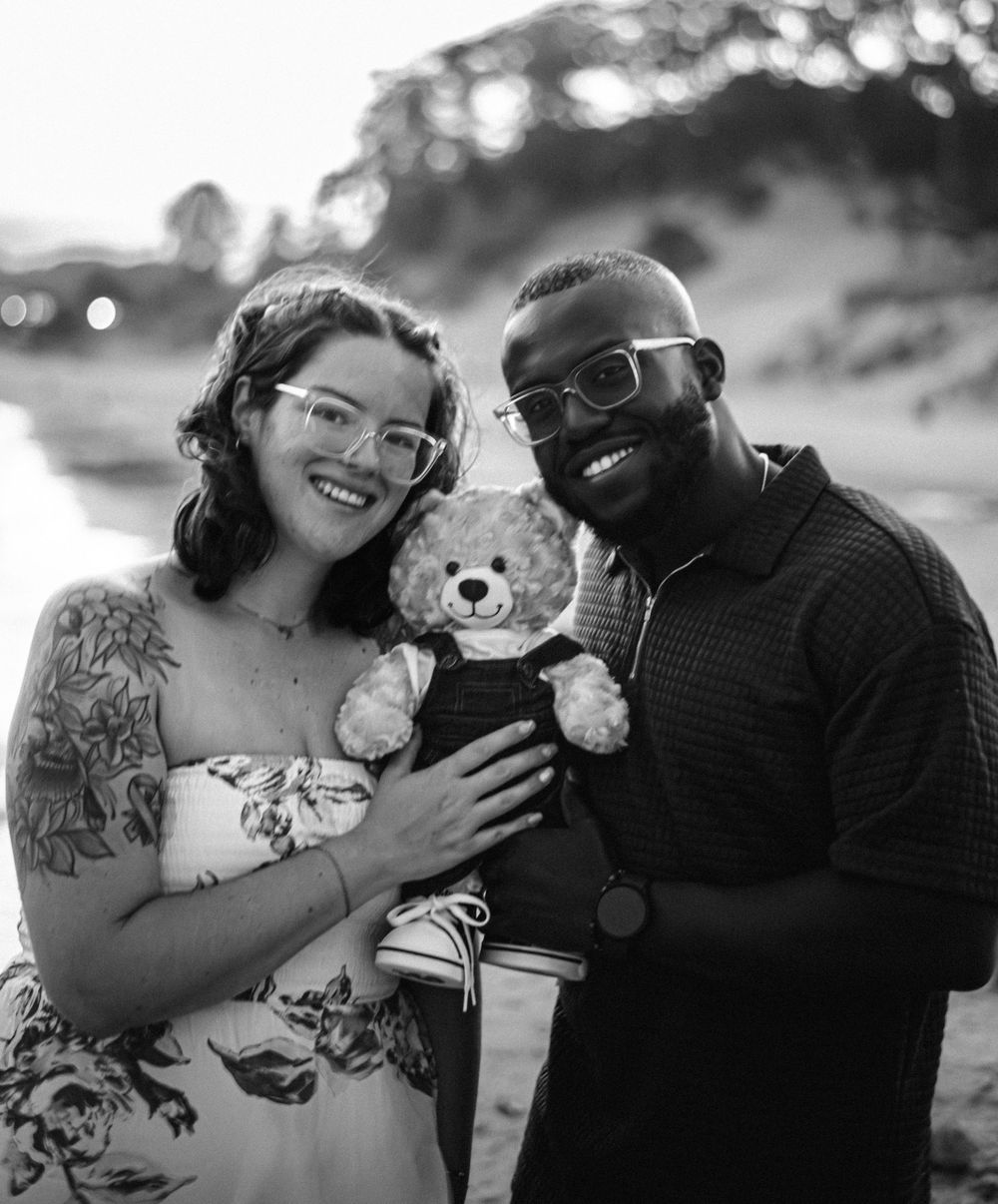 A couple poses with a teddy bear during a beach photoshoot at sunset.