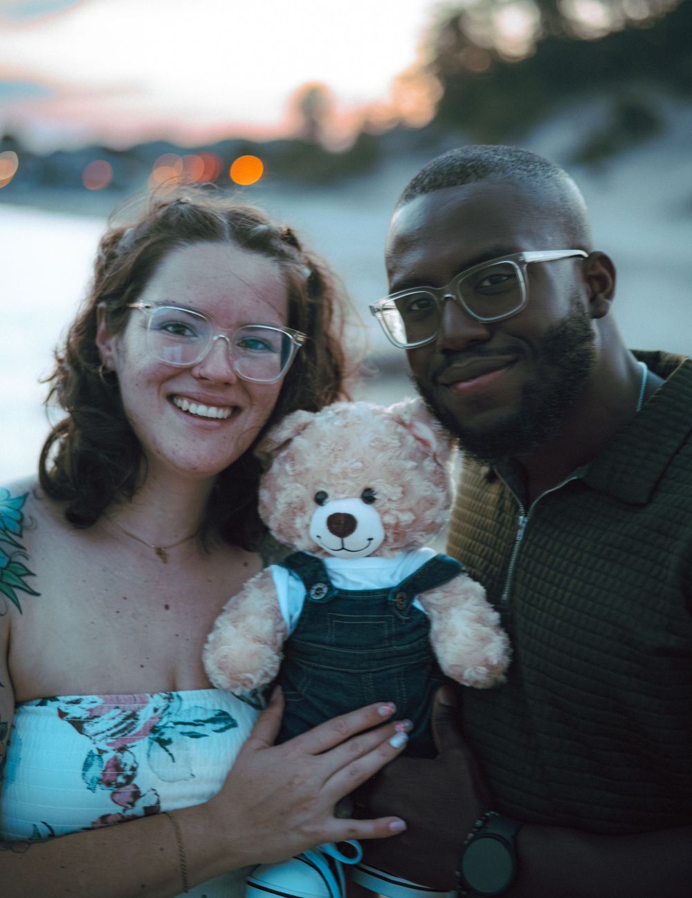 A couple shares joyful moments with a plush teddy bear during their beach announcement photoshoot.