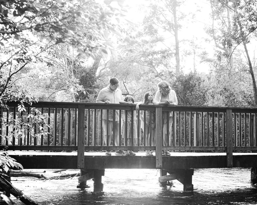 Black and white photo of a family standing on a wooden bridge surrounded by lush trees and a stream below.