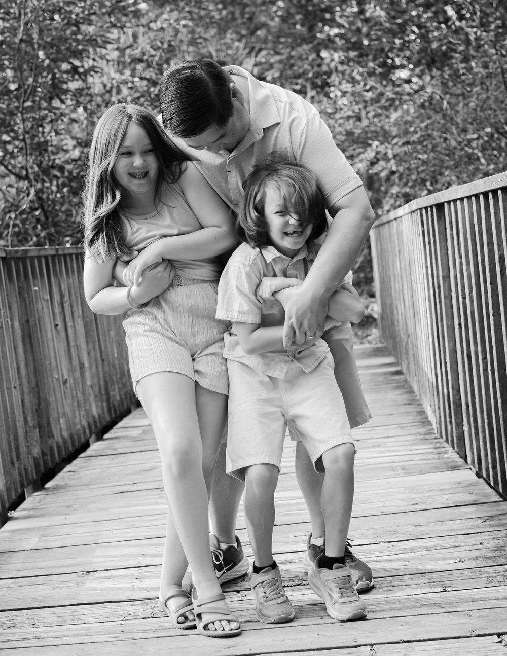 Black and white photo of a father playfully hugging his two laughing children on a wooden bridge outdoors.