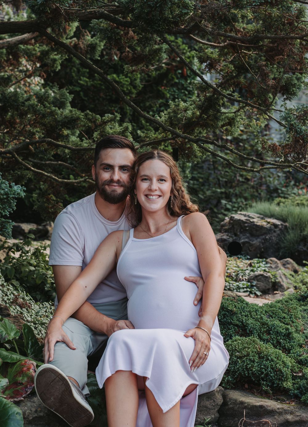 A couple in white outfits poses together on stone steps surrounded by lush greenery.
