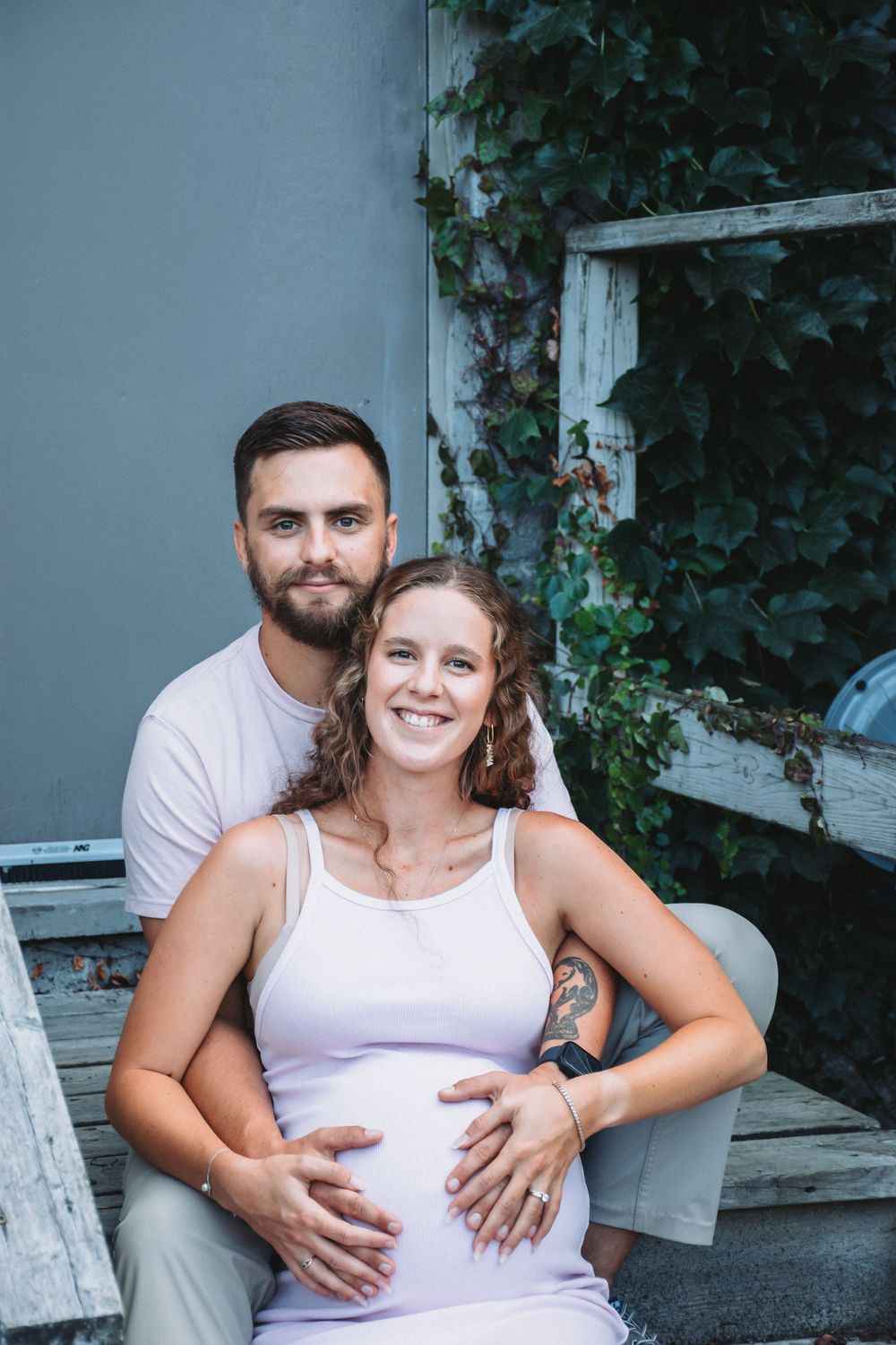 Outdoor maternity photo session with couple sitting on wooden bench surrounded by ivy.