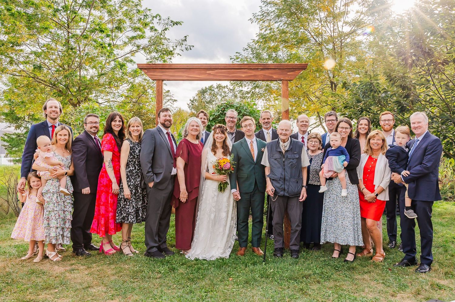 Wedding party gathered outdoors in front of a wooden archway during sunset with trees and natural scenery in background.