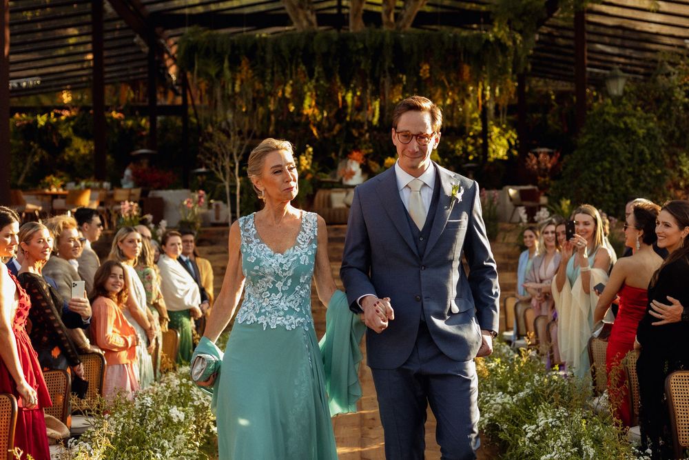 Wedding couple walks down floral-lined aisle in rustic venue with guests applauding.