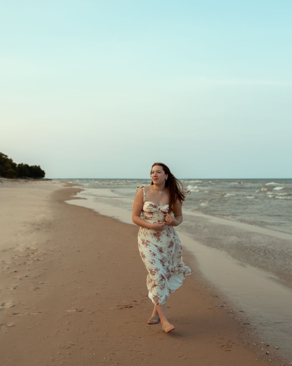 A person in a floral dress dances gracefully along a serene beach shoreline at sunset.